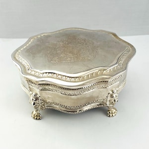 May include: A silver-colored, ornate jewelry box with a scalloped edge and lion's paw feet. The lid features an engraved crest design. The box is likely made of metal and sits on a white surface.