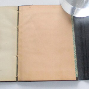 May include: A handmade journal with cream and tan pages, bound with black thread. The journal has a black, textured cover with a unique shape. A magnifying glass rests on the journal.