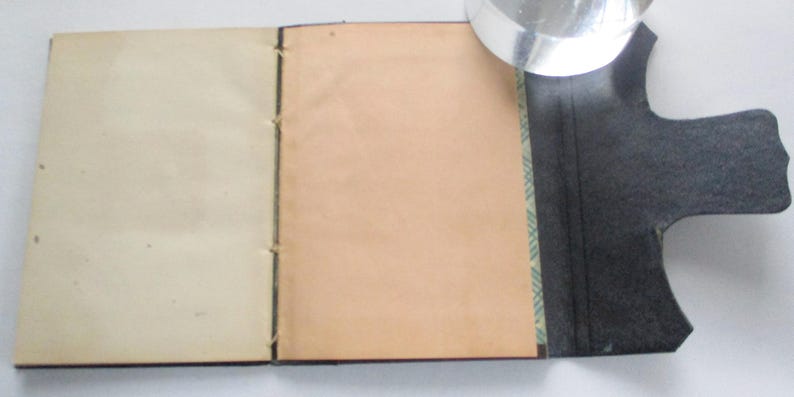 May include: A handmade journal with cream and tan pages, bound with black thread. The journal has a black, textured cover with a unique shape. A magnifying glass rests on the journal.