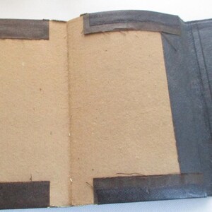 May include: An open vintage book, showing a tan interior and a dark grey exterior. The cover has brown fabric strips for reinforcement and a patterned green and white edge. The book is open, revealing the inside pages.