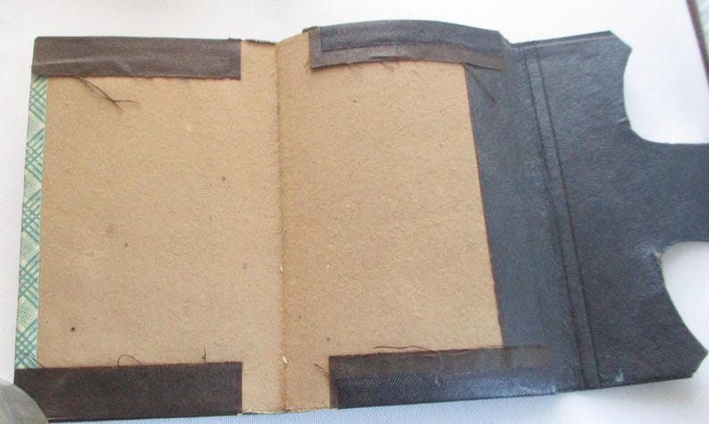 May include: An open vintage book, showing a tan interior and a dark grey exterior. The cover has brown fabric strips for reinforcement and a patterned green and white edge. The book is open, revealing the inside pages.