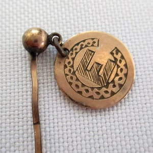 May include: A gold-toned metal pin with a round, engraved head featuring a stylized letter "E" surrounded by a decorative border. The pin has a long, thin stem.