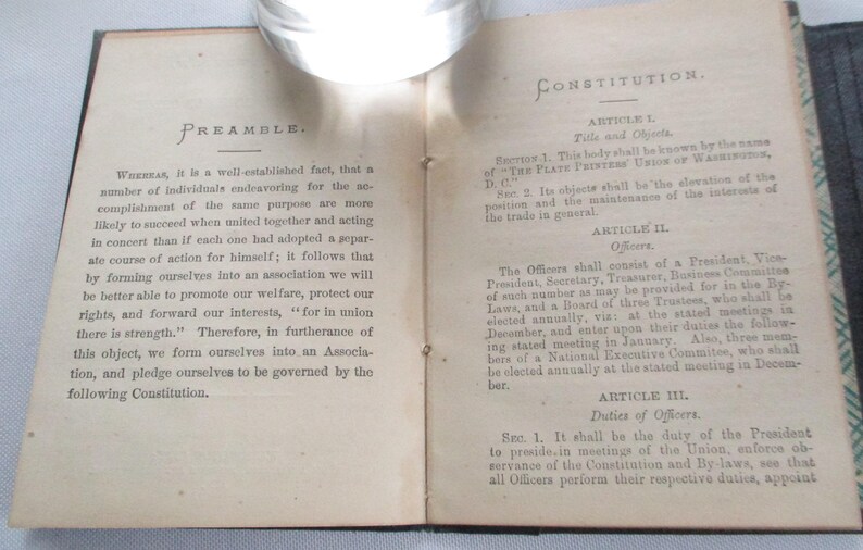 May include: An open book with aged, off-white pages. The left page reads "PREAMBLE" with text discussing the formation of an association. The right page displays "CONSTITUTION" and article titles. The book is bound in a dark cover.