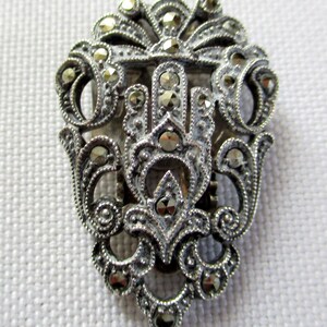 Puede incluir: Un broche plateado con un dise&ntilde;o detallado que presenta un motivo central de una mano estilizada rodeada de intrincados patrones florales. El broche est&aacute; adornado con numerosas peque&ntilde;as piedras de marcasita brillantes.