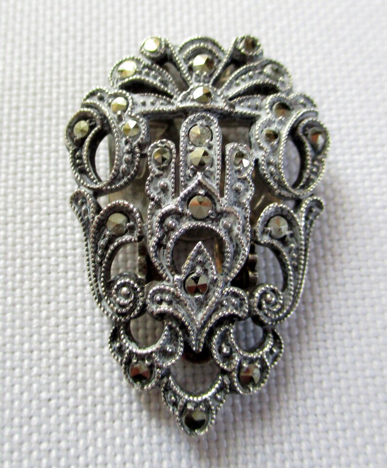 Puede incluir: Un broche plateado con un dise&ntilde;o detallado que presenta un motivo central de una mano estilizada rodeada de intrincados patrones florales. El broche est&aacute; adornado con numerosas peque&ntilde;as piedras de marcasita brillantes.