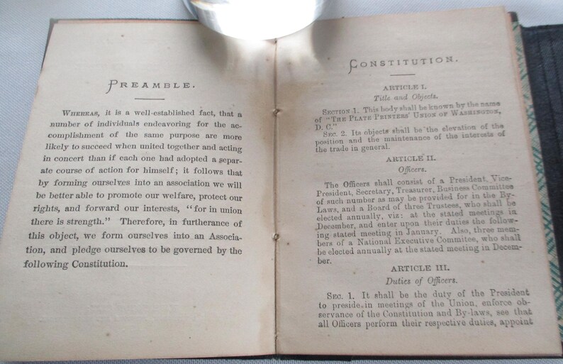 May include: An open book displaying text on aged paper. The left page is headed "PREAMBLE" and the right page "CONSTITUTION." The text is in black ink. The book is open at the centre, showing the binding.