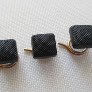 May include: Three black square buttons with a textured surface are displayed. Each button is attached to a gold-colored metal pin. The buttons are arranged on a white, textured surface.