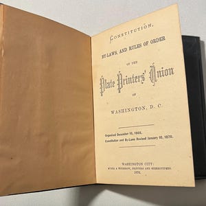 May include: An open book with a black cover and aged, tan pages. The right page features the title "Constitution, By-Laws, and Rules of Order of the Plate Printers' Union of Washington, D.C."