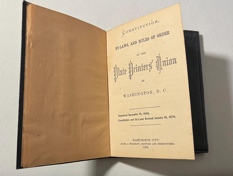 May include: An open book with a black cover and aged, tan pages. The right page features the title "Constitution, By-Laws, and Rules of Order of the Plate Printers' Union of Washington, D.C."