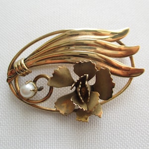 May include: A vintage gold-toned brooch featuring a floral design with a pearl accent. The brooch has a stylized flower and leaf arrangement within an oval frame. The pearl is small and round.