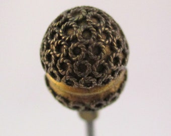 May include: A gold-toned pin with a round, intricate, filigree design. The pin is attached to a thin, silver-colored metal pin.
