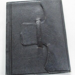 May include: A black, rectangular, leather-bound book with a textured surface. The book has a strap closure and a decorative flap. The cover is a solid black colour.