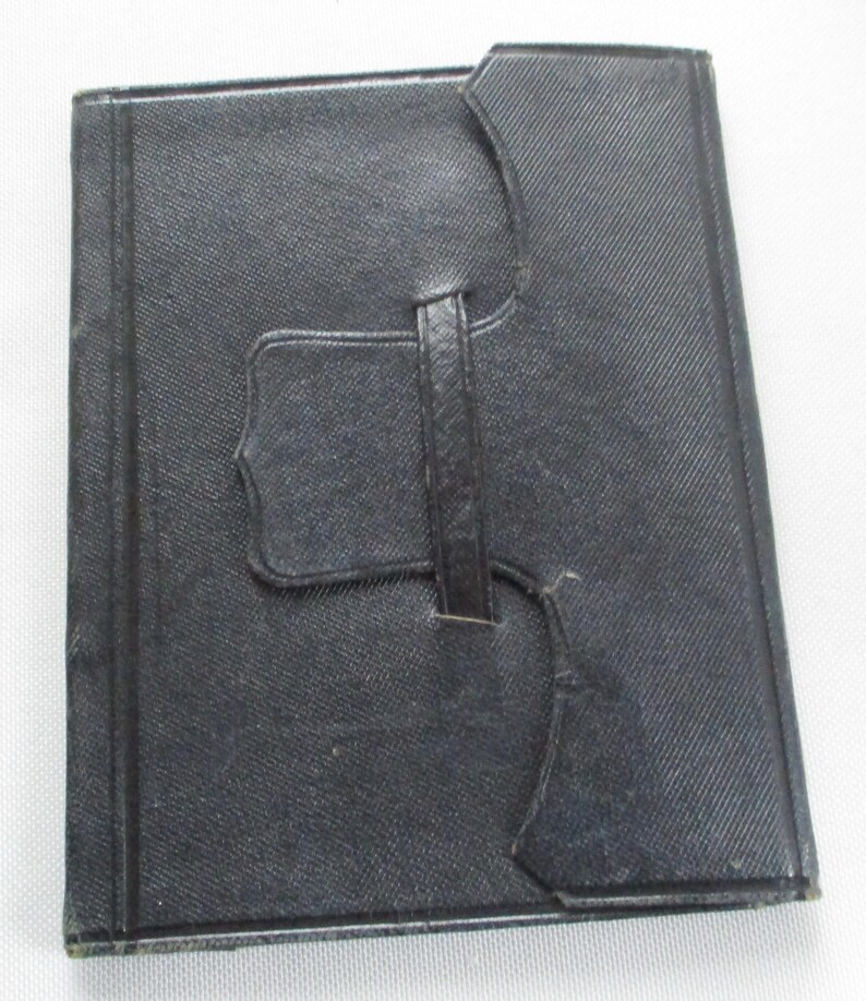 May include: A black, rectangular, leather-bound book with a textured surface. The book has a strap closure and a decorative flap. The cover is a solid black colour.