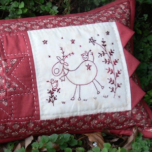 Op de afbeelding: Rechthoekig decoratief kussen met een rood-wit patchwork ontwerp. Het kussen heeft een centraal paneel met een gestikte ster en twee witte panelen met geborduurde rendieren en vogelmotieven. De randen zijn afgewerkt met een bloemenpatroon.