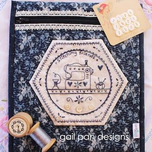 May include: A blue fabric zippered pouch with a white lace trim. The pouch features a hexagonal patch with an embroidered design of a sewing machine, thread, and needles. The text "Stitching friends" is embroidered on the patch. The pouch also has a label that reads "gail pan designs".
