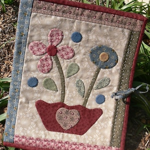 Può includere: Un arazzo da parete trapuntato con un motivo floreale in tessuto rosa, rosso, blu e verde. I fiori sono in un vaso rosso con un design a forma di cuore. L'arazzo ha uno sfondo a motivi marroni e bianchi.