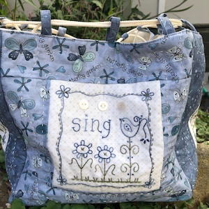 Op de afbeelding: Een blauwe gewatteerde draagtas met een vlinder- en bloemenpatroon. De tas heeft een voorvak met het woord "sing" geborduurd boven een vogel en bloemen. De handvatten zijn van blauwe stof en een wit touw.