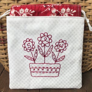 Puede incluir: Bolsa de cordón blanca con un patrón floral rojo en el interior. La parte delantera de la bolsa tiene un diseño bordado rojo de tres flores en una maceta con las letras S, W y & en el centro de cada flor.