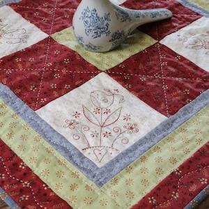 May include: A white quilted table runner with a red, green, and blue floral design. The runner has a decorative border with a blue and white bird in the center.