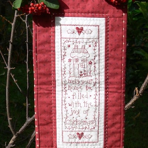 Può includere: Un banner natalizio rosso e bianco con un disegno a casa ricamata e la scritta "May your home be filled with the joy of Christmas". Il banner è adornato con bacche rosse e una parte superiore in metallo decorativa.