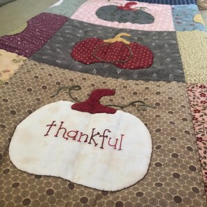 May include: A patchwork quilt with a brown background and a white pumpkin with the word "thankful" embroidered on it. The quilt features a variety of colors and patterns, including red, green, and blue.