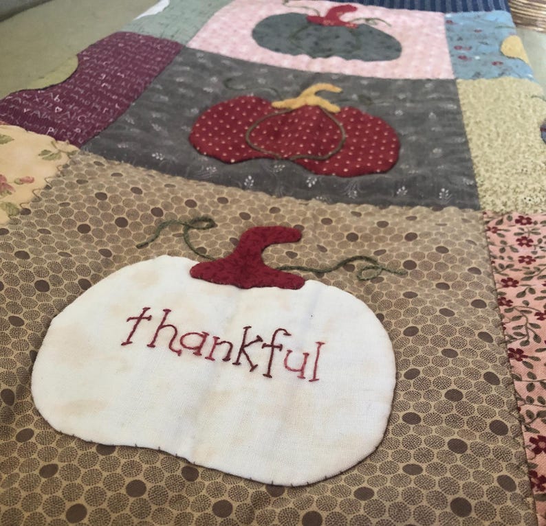 May include: A patchwork quilt with a brown background and a white pumpkin with the word "thankful" embroidered on it. The quilt features a variety of colors and patterns, including red, green, and blue.
