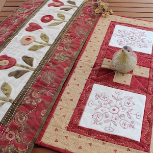 Może przedstawiać: Dwa ręcznie robione pikowane bieżniki na stół. Jeden jest prostokątny z kwiatowymi aplikacjami na białym środku, obramowany czerwoną i brązową wzorzystą tkaniną. Drugi jest kwadratowy z haftowanymi wzorami płatków śniegu na białych kwadratach, na czerwonym i beżowym tle.