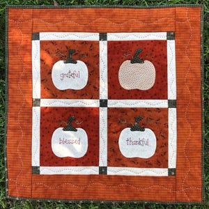 May include: A white quilted wall hanging with four white pumpkins. Each pumpkin has a different word embroidered on it: grateful, blessed, thankful, and happy. The background is a brown and orange fabric with a white polka dot pattern.