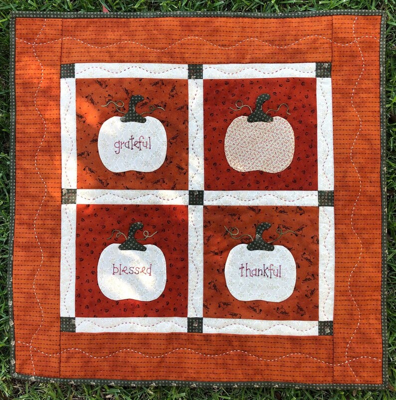 May include: A white quilted wall hanging with four white pumpkins. Each pumpkin has a different word embroidered on it: grateful, blessed, thankful, and happy. The background is a brown and orange fabric with a white polka dot pattern.