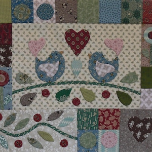 Può includere: Un quadrato di patchwork con due uccelli blu con cuori rosa, un cuore rosso e varie foglie e bacche. Il disegno è su uno sfondo color crema con motivi floreali, circondato da quadrati di tessuto fantasia.