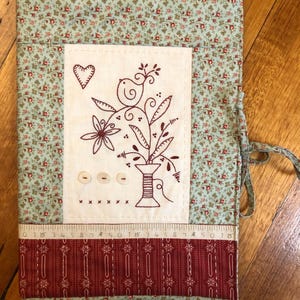 May include: A green and red fabric needle case with a white embroidered design of a bird, flowers, and a spool of thread. The case has a drawstring closure and a ruler printed on the bottom.
