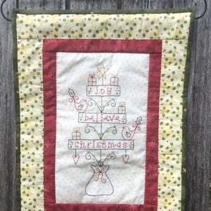 May include: A quilted Christmas wall hanging with a festive tree design. The tree is adorned with gifts, stars, and the words "joy", "believe", and "christmas". The border is red and green, with a decorative metal hanger.