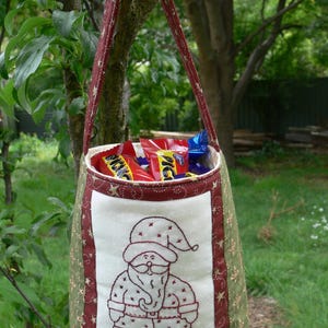 May include: A green and red fabric bag with a white panel featuring an embroidered Santa Claus. The bag is filled with chocolate bars.