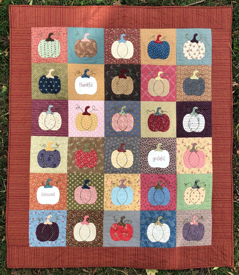 May include: A patchwork quilt with a red border and a pattern of pumpkins in various colors and designs. The quilt has the words "thankful", "grateful", and "blessed" stitched into it.