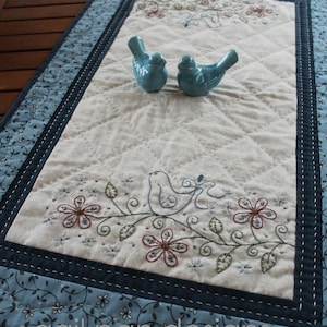 May include: A cream-coloured quilted table runner with embroidered flowers and a bird design. Two small blue ceramic bird figurines are placed on the runner. The border is a light blue floral pattern with a dark blue trim.