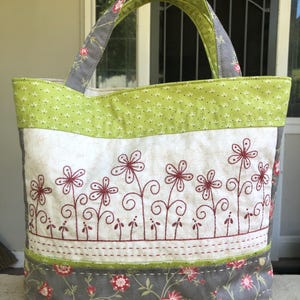 Pode incluir: Uma sacola tote feita à mão com um design floral. A sacola apresenta um painel central branco com flores vermelhas bordadas, emoldurado por tecido floral verde e cinza. As alças são cinza com detalhes florais vermelhos.