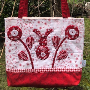 Puede incluir: Bolso tote acolchado con fondo blanco con diseño floral y conejito rojo. El bolso tiene asas rojas y un ribete de cuadros rojos y blancos. La parte inferior del bolso es de color rojo liso.