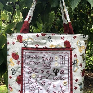 May include: A white tote bag with a sewing-themed pattern. The bag features a red border and handles, with embroidered details including the words "create buttons sew needles". The bag has a strawberry, scissors, and spool design.