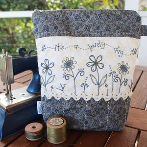 Op de afbeelding: Een grijze en witte stoffen etui met een rits en een wit stoffen paneel met geborduurde bloemen en de tekst "it's a lovely day".