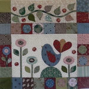 Puede incluir: Un edredón de retazos con un diseño central de un pájaro azul con detalles en rojo y marrón, rodeado de flores estilizadas y hojas verdes. El edredón tiene un fondo beige con varios cuadrados estampados.