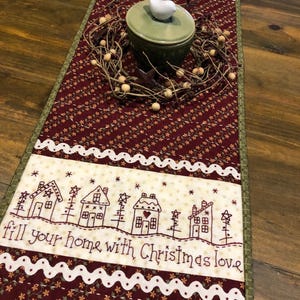 May include: A red and green quilted table runner with a white embroidered border and the text "fill your home with Christmas love". The runner features a design of small houses and trees. A white bird sits on top of a green ceramic container.