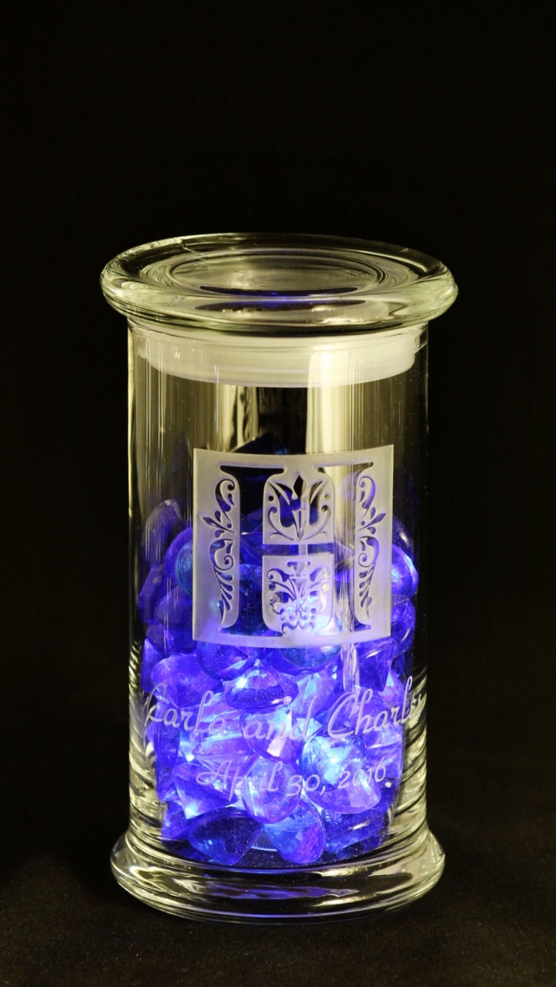May include: A clear glass jar with a lid, filled with blue and purple glass gems. The jar is engraved with the letters "T & C" and the text "Tara and Charles April 30, 2016".