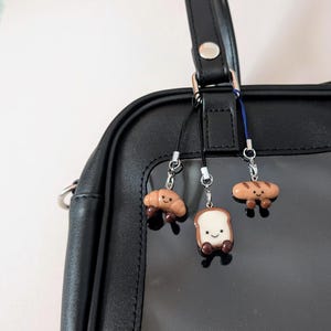 May include: Three food-themed charms suspended from a black bag. The charms depict a croissant, a slice of bread, and a loaf of bread, each with smiling faces and tiny legs. The charms are brown and white.