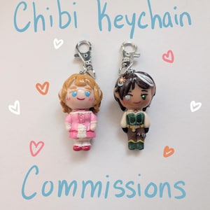 May include: Two chibi character keychains with silver lobster clasps. One figure has light brown hair and a pink dress. The other has dark hair with a white streak and a green and brown outfit. The text "Chibi Keychain Commissions" is visible.