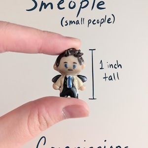 May include: A miniature figurine titled Smeople (small people) Commissions, depicting a character with dark hair, a beige trench coat, and a blue tie. The figurine is 1 inch tall, featuring a glossy finish and detailed facial features, presented as a custom commission piece.