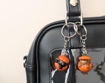 May include: Two Halloween-themed keyrings suspended from a black handbag. One shows a black cat in an orange pumpkin. The other has a brown bear wearing an orange pumpkin hat. Both have silver chains, perfect for spooky season.