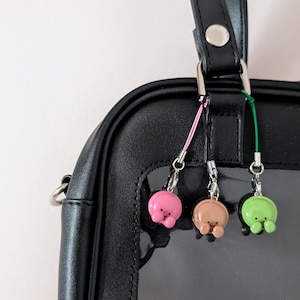 Puede incluir: Primer plano de un bolso negro con tres colgantes de colores colgando del asa. Los colgantes son rosa, marrón y verde, cada uno con un cuerpo pequeño y redondo y patas diminutas. Los colgantes están sujetos al bolso con cierres plateados.