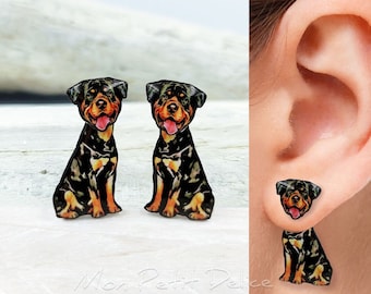 May include: A pair of stud earrings featuring a black and brown Rottweiler dog design. The earrings are shown on a white surface and in a model's ear.