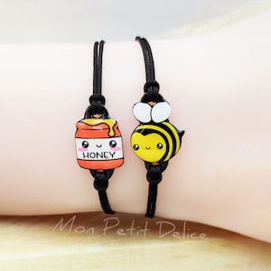 May include: Two black friendship bracelets with cute charms. One charm is a honey jar with a smiling face and the word "HONEY" on it. The other charm is a cartoon bee with a smiling face.