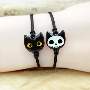 May include: Two black cord bracelets with charms. One charm is a black cat with yellow eyes. The other charm is a white skull with black details.